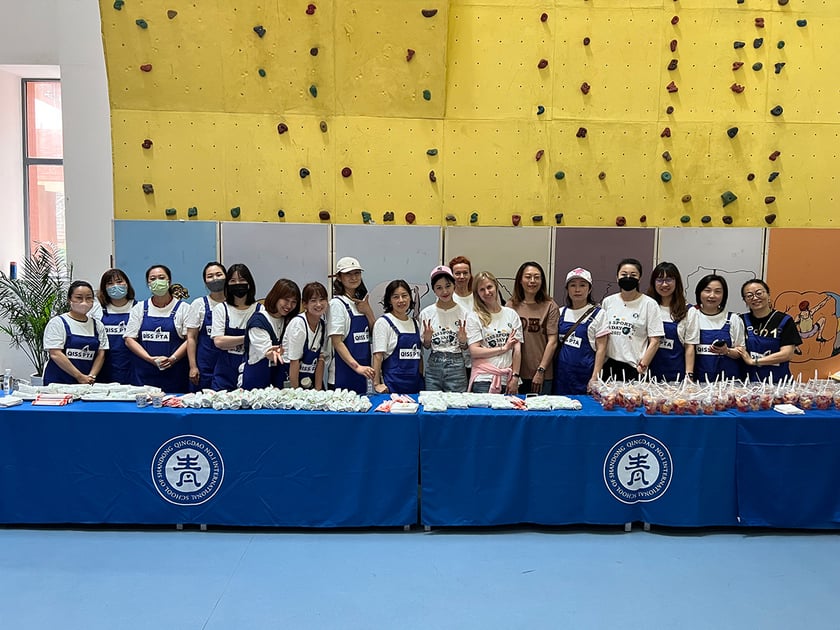 Extracurriculars That Strengthen College Applications 3 QISS students and staff in blue aprons run a school fundraiser booth, showcasing community service as a standout extracurricular on college applications