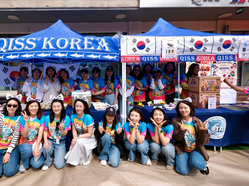 International Day at School: What Parents Should Know 3 Parents in matching tie-dye shirts gather in front of the QISS Korea stall at International Day